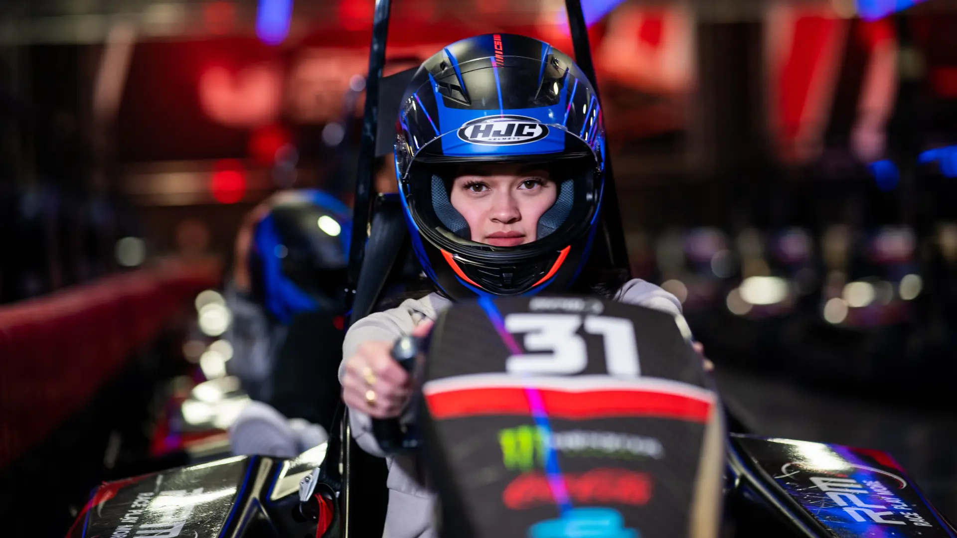 A person wearing a black helmet with a blue visor is seated in a go-kart, gripping the steering wheel. The go-kart, marked with the number 31, is set against a blurred backdrop of bright red and blue lights, evoking the thrilling atmosphere of an indoor race at RPM Raceway.