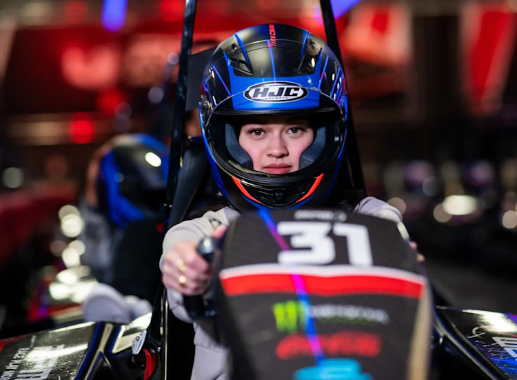 A person wearing a black helmet with a blue visor is seated in a go-kart, gripping the steering wheel. The go-kart, marked with the number 31, is set against a blurred backdrop of bright red and blue lights, evoking the thrilling atmosphere of an indoor race at RPM Raceway.