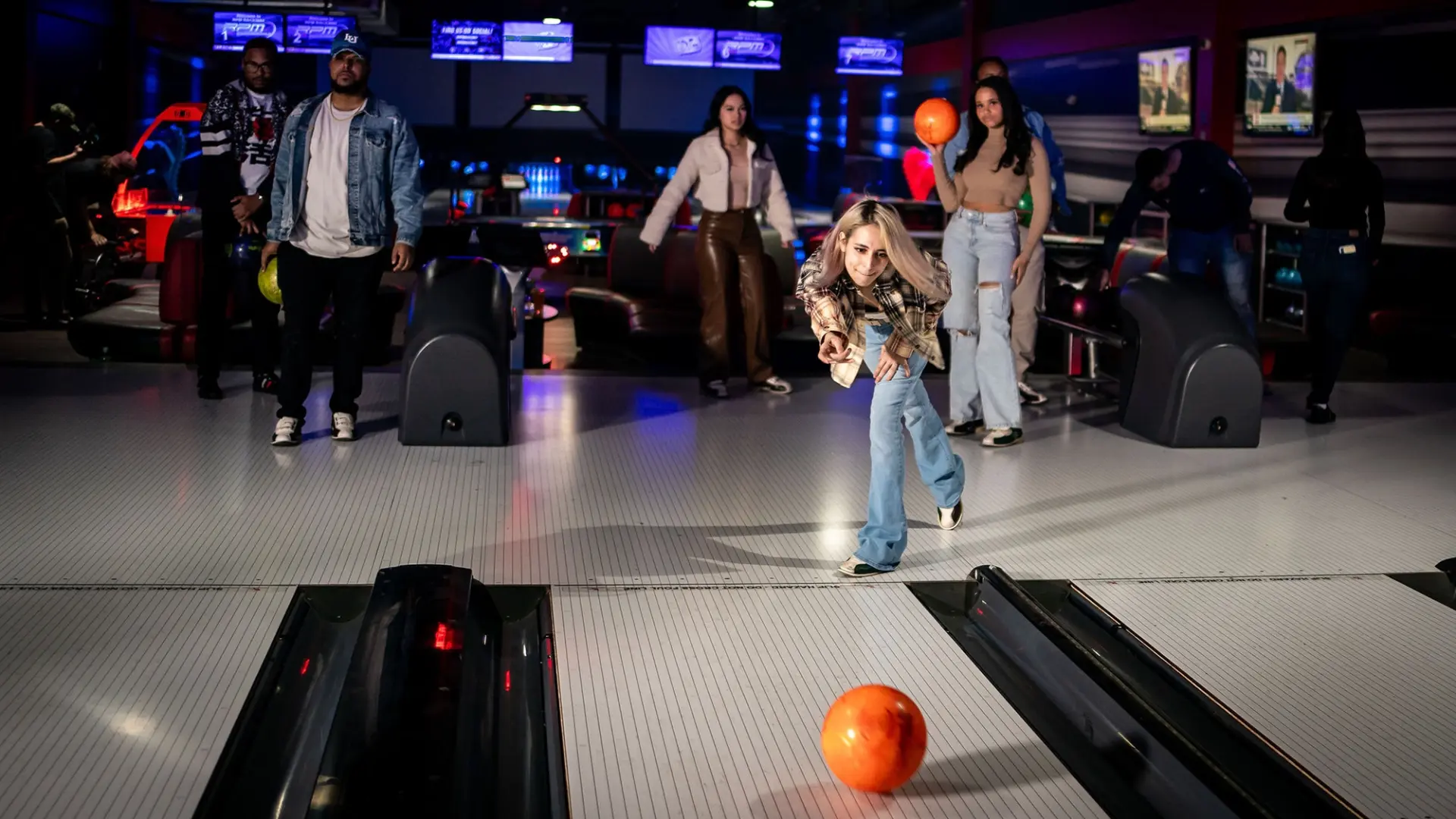 A young girl throws a bowling ball down a lane in a bowling alley, while four other people watch. The alley is brightly lit with colorful lights and screens displaying scores in the background. The atmosphere appears lively and engaging.