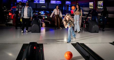 A young girl throws a bowling ball down a lane in a bowling alley, while four other people watch. The alley is brightly lit with colorful lights and screens displaying scores in the background. The atmosphere appears lively and engaging.