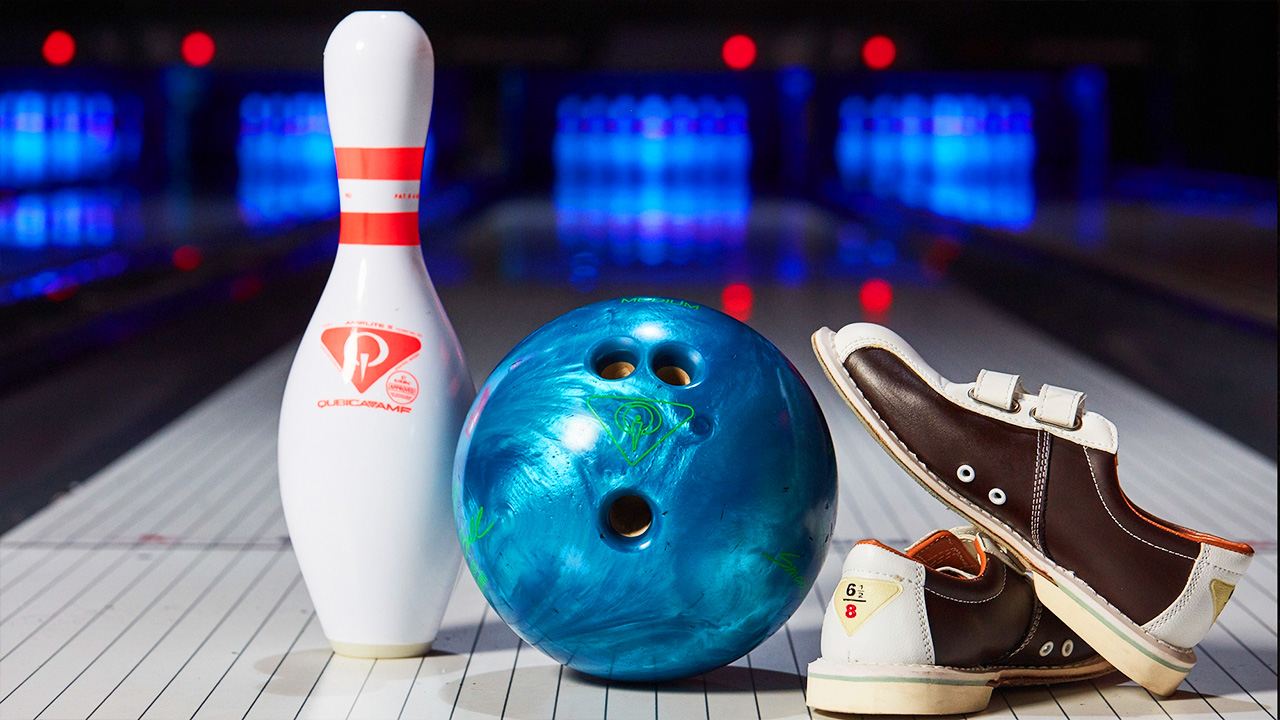 A young girl throws a bowling ball down a lane in a bowling alley, while four other people watch. The alley is brightly lit with colorful lights and screens displaying scores in the background. The atmosphere appears lively and engaging.