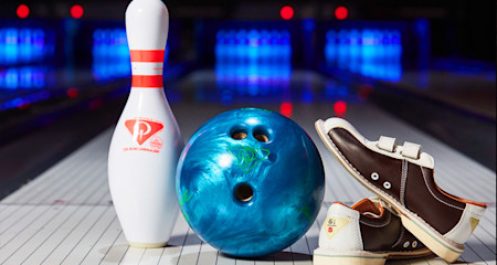 A young girl throws a bowling ball down a lane in a bowling alley, while four other people watch. The alley is brightly lit with colorful lights and screens displaying scores in the background. The atmosphere appears lively and engaging.
