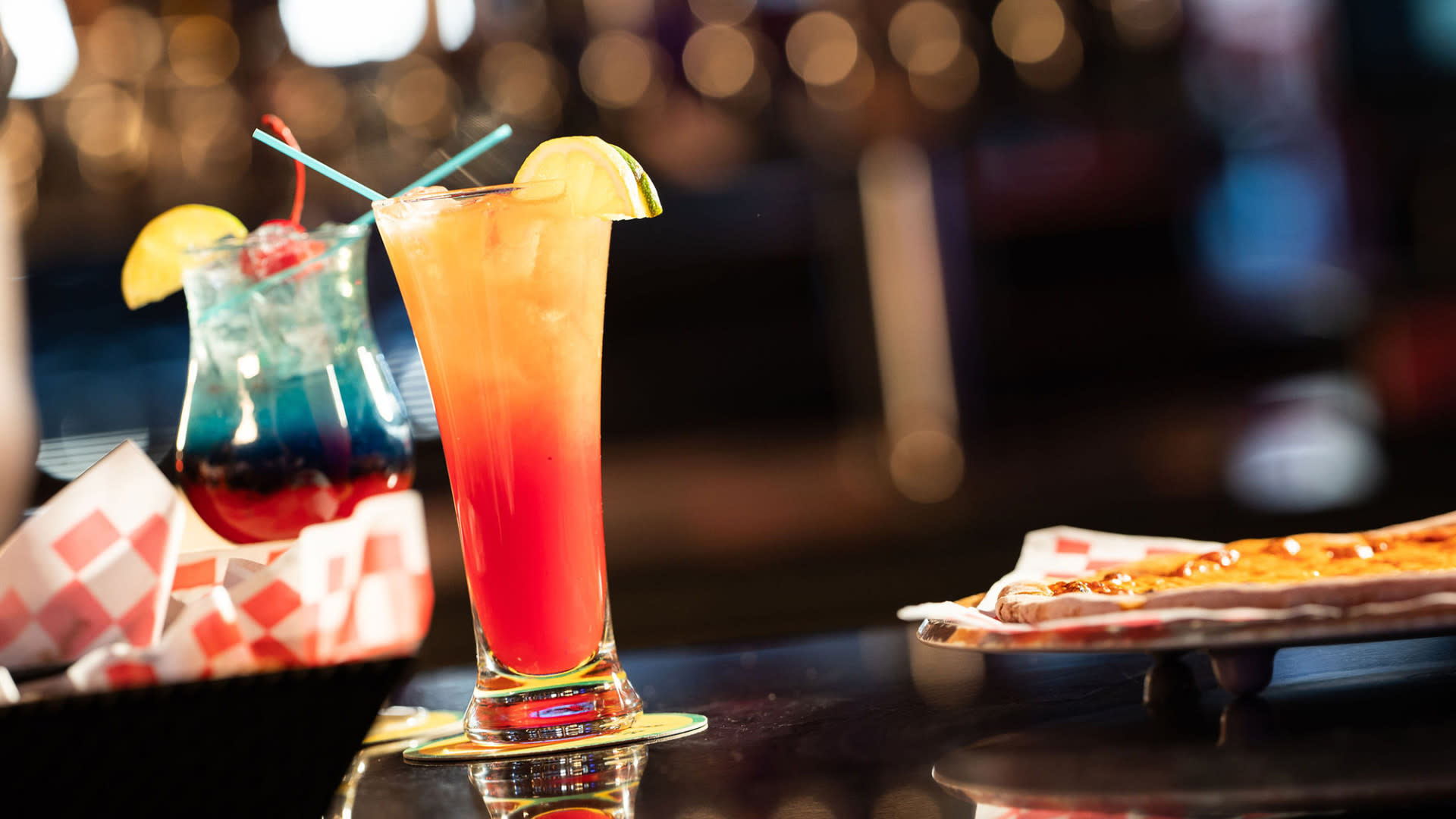 Two colorful cocktails sit on a bar counter. The drink on the left is blue with a cherry garnish and a straw, while the drink on the right is a gradient of red, orange, and yellow with a lime wedge. A plate with food is partially visible on the right side.