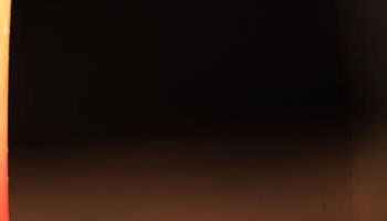 Two colorful cocktails sit on a bar counter. The drink on the left is blue with a cherry garnish and a straw, while the drink on the right is a gradient of red, orange, and yellow with a lime wedge. A plate with food is partially visible on the right side.