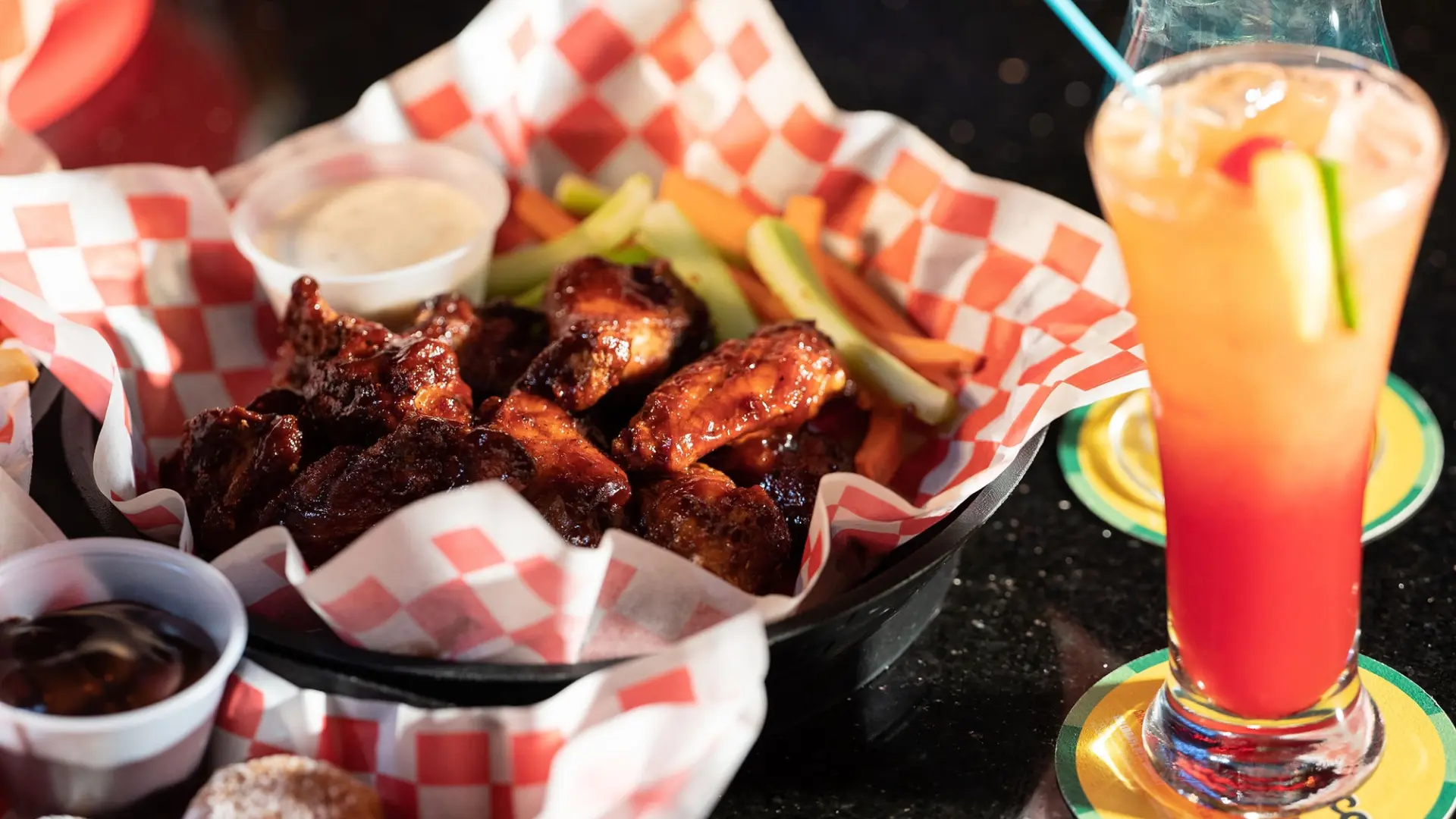 A serving of sauced chicken wings is placed in a basket lined with red and white checkered paper, accompanied by celery sticks and a small container of dipping sauce. Perfect for enjoying at a lively sports bar, a fruity cocktail with a straw and garnish sits on a coaster to the right.