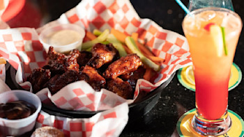 A serving of sauced chicken wings is placed in a basket lined with red and white checkered paper, accompanied by celery sticks and a small container of dipping sauce. Perfect for enjoying at a lively sports bar, a fruity cocktail with a straw and garnish sits on a coaster to the right.