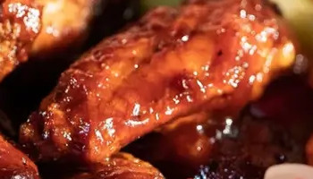 A serving of sauced chicken wings is placed in a basket lined with red and white checkered paper, accompanied by celery sticks and a small container of dipping sauce. Perfect for enjoying at a lively sports bar, a fruity cocktail with a straw and garnish sits on a coaster to the right.