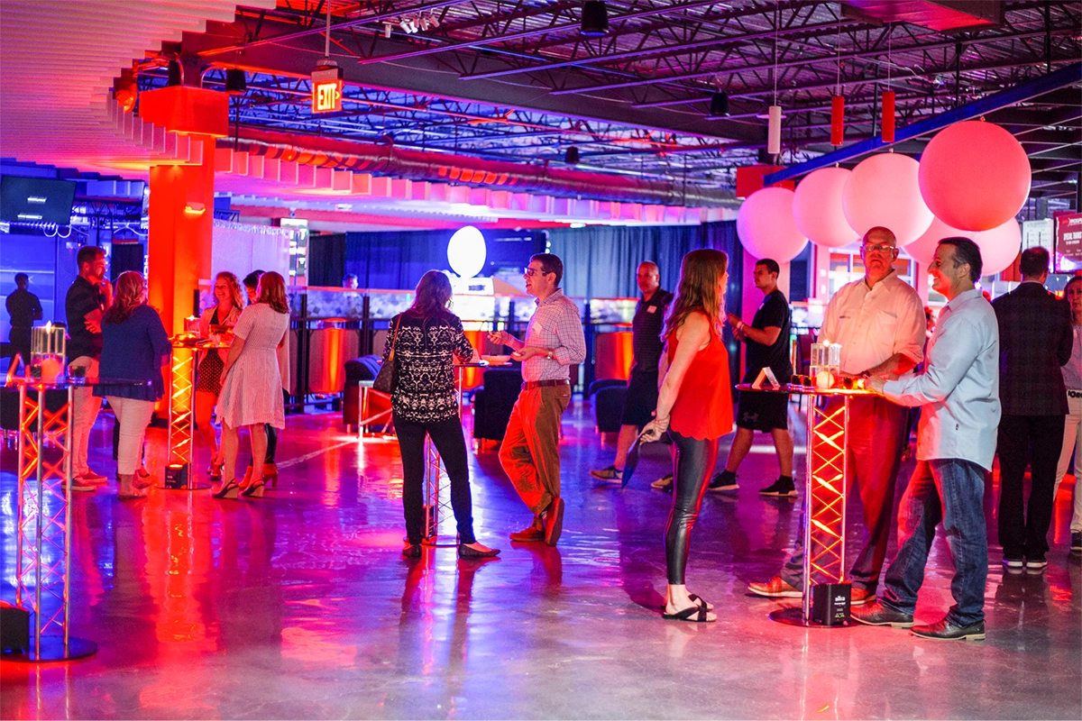 People are socializing at an indoor event with neon lighting. The room is decorated with large white balloons and illuminated furniture. Several groups are standing or sitting at high tables with drinks. The atmosphere is lively and colorful.