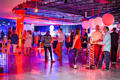 People are socializing at an indoor event with neon lighting. The room is decorated with large white balloons and illuminated furniture. Several groups are standing or sitting at high tables with drinks. The atmosphere is lively and colorful.