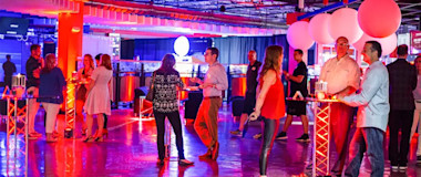 People are socializing at an indoor event with neon lighting. The room is decorated with large white balloons and illuminated furniture. Several groups are standing or sitting at high tables with drinks. The atmosphere is lively and colorful.