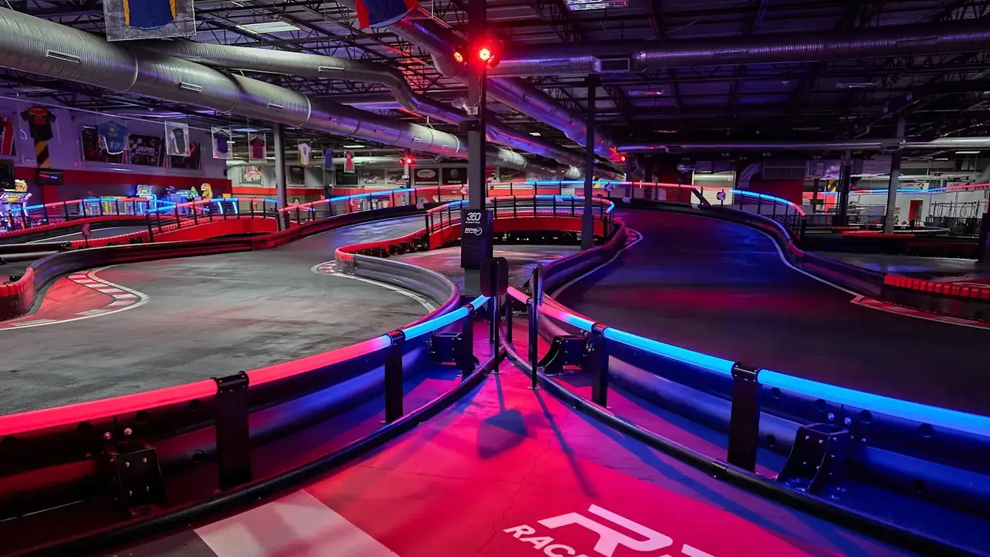 Indoor go-kart track with red and blue neon lighting. The track features sharp turns and barriers with a smooth racing surface. Overhead pipes and industrial lighting fill the ceiling. The track is empty, highlighting its futuristic, high-tech design.