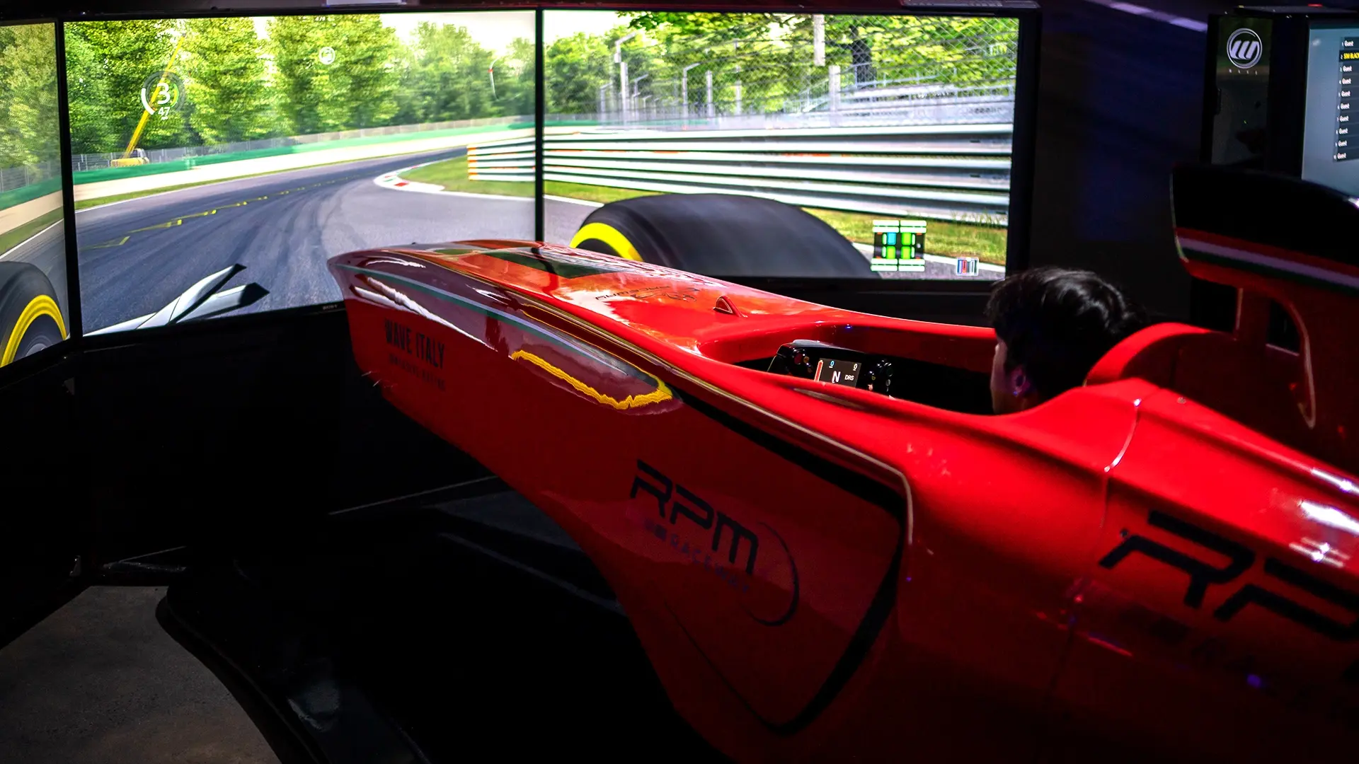 A person is sitting in a red racing car simulator cockpit, with a large curved screen displaying a realistic race track and surrounding greenery. The go-kart themed simulator is detailed with various logos and text. The scene is indoors, with dim lighting focused on this thrilling entertainment setup.