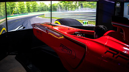 A person is sitting in a red racing car simulator cockpit, with a large curved screen displaying a realistic race track and surrounding greenery. The go-kart themed simulator is detailed with various logos and text. The scene is indoors, with dim lighting focused on this thrilling entertainment setup.