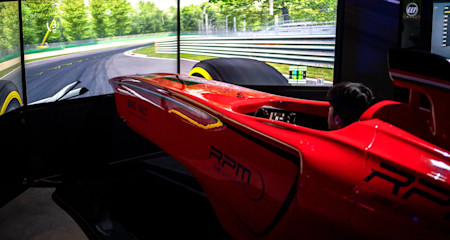 A person is sitting in a red racing car simulator cockpit, with a large curved screen displaying a realistic race track and surrounding greenery. The go-kart themed simulator is detailed with various logos and text. The scene is indoors, with dim lighting focused on this thrilling entertainment setup.