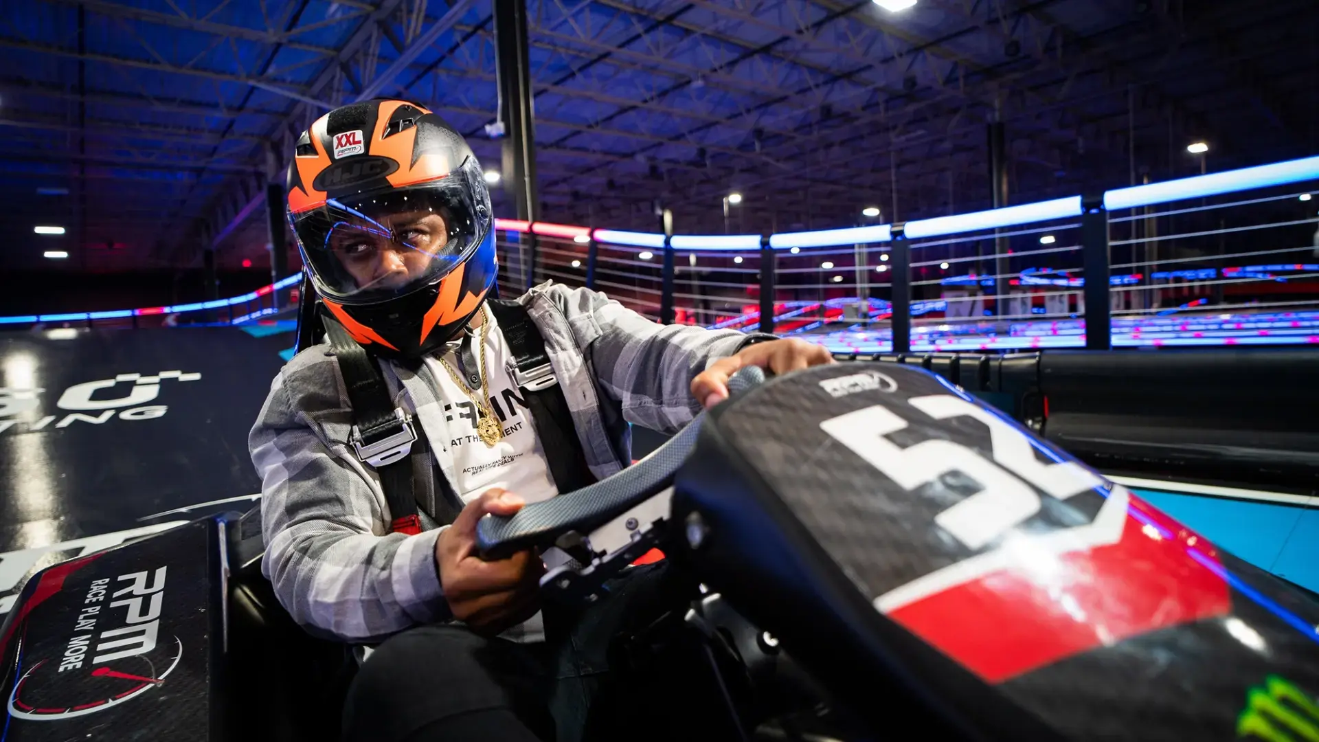 A person wearing an orange and black helmet with a clear visor is driving an RPM go-kart. The go-kart has a black body with the number 57 on the front. They are wearing a gray plaid jacket and a harness. Blurred neon lights create a vibrant background on the indoor race track.