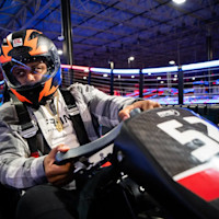 A person wearing an orange and black helmet with a clear visor is driving an RPM go-kart. The go-kart has a black body with the number 57 on the front. They are wearing a gray plaid jacket and a harness. Blurred neon lights create a vibrant background on the indoor race track.