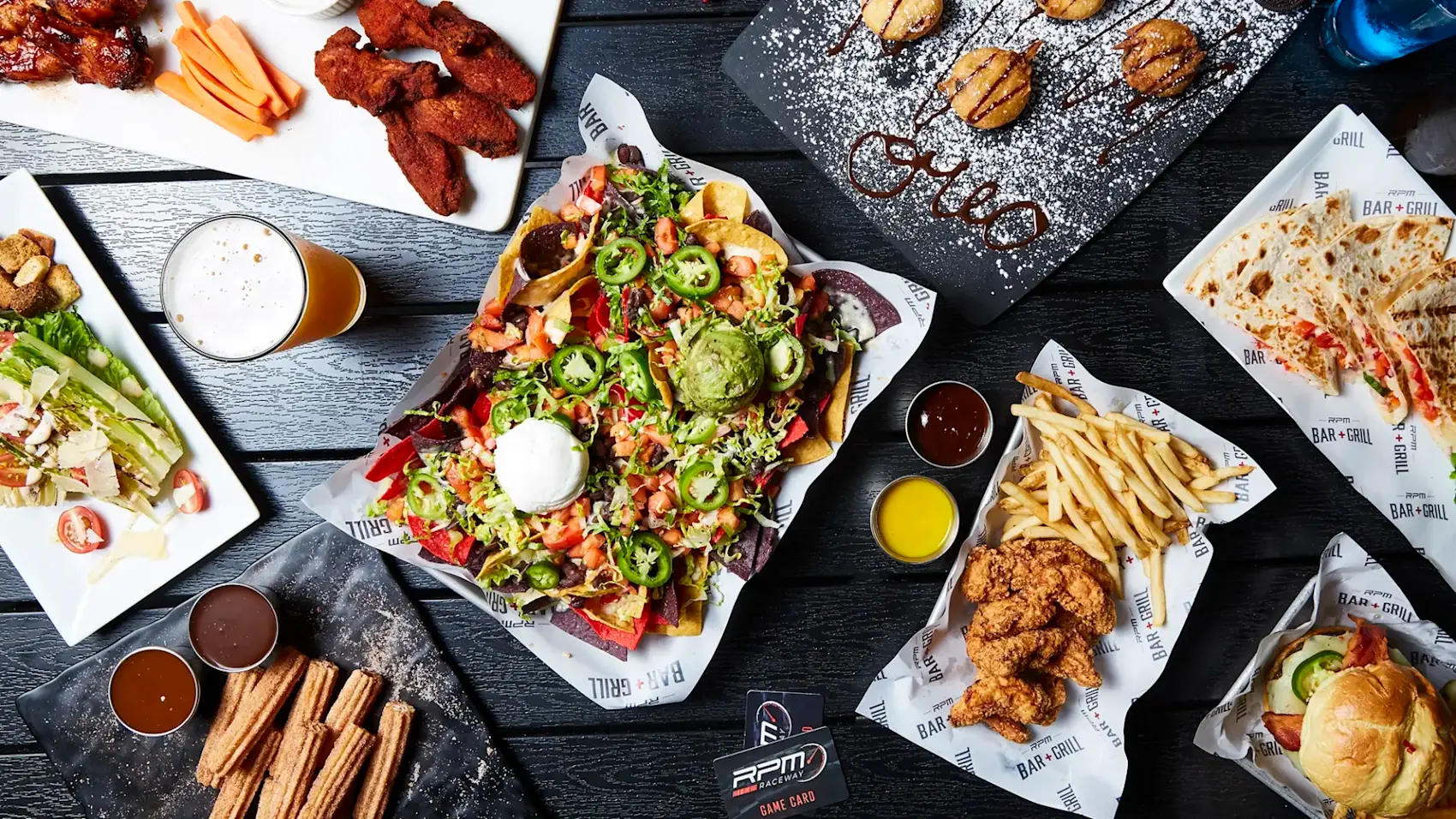 A flat lay photo of a table filled with various dishes, including nachos topped with guacamole and sour cream, fried chicken, fries, buffalo wings, salad, churros, and dessert. Drinks are also visible, along with sauce dips served on a dark table.