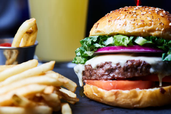 A close-up of a juicy cheeseburger with lettuce, tomato, onions, and melted cheese on a sesame seed bun, accompanied by a side of crispy French fries. In the background, there is a blurred drink.