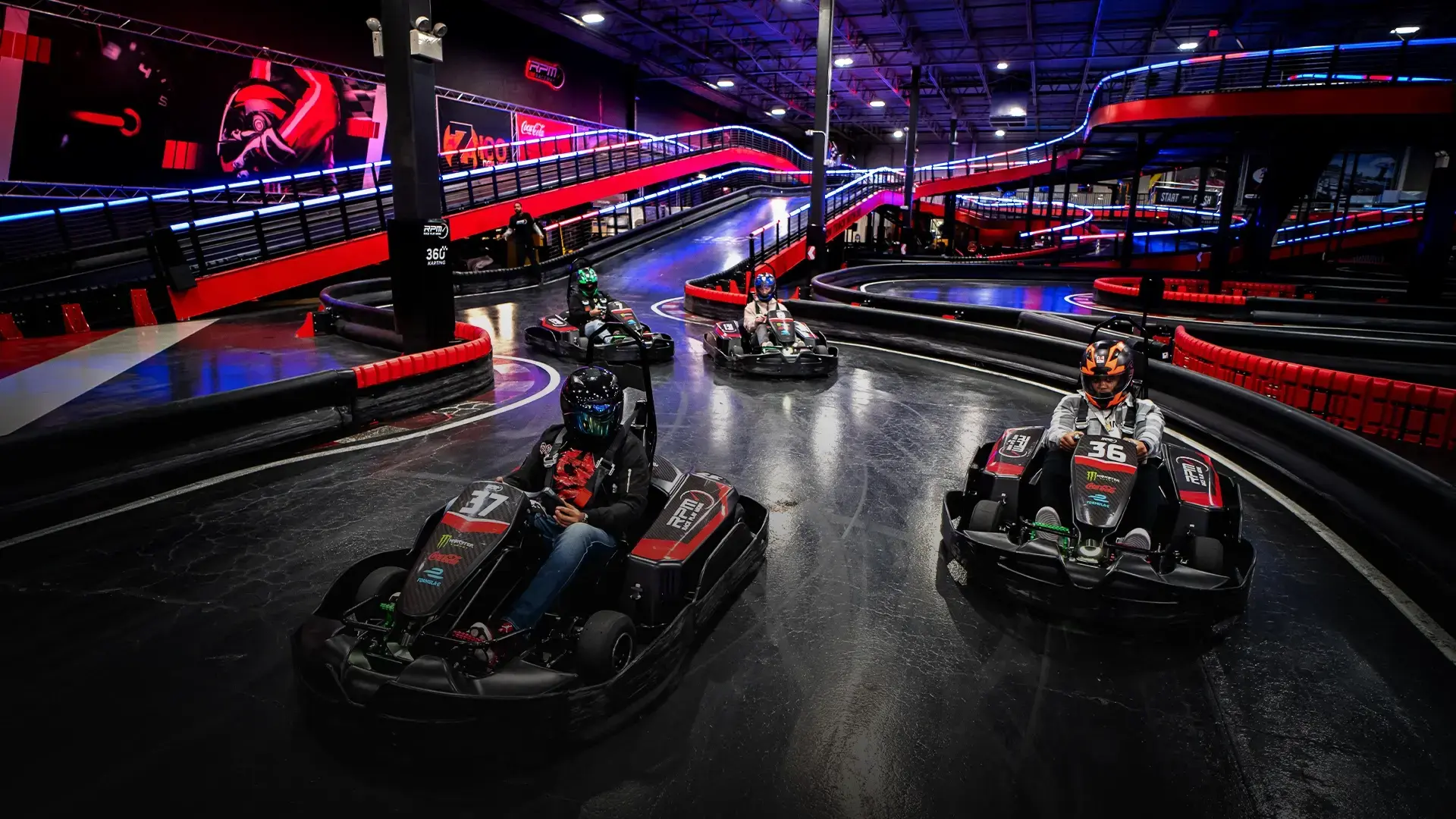 A group of go-kart racers navigate a track with neon lights and red barriers inside an indoor racing facility. The track is winding, with an elevated section visible in the background. The racers wear helmets and are in individual go-karts marked with numbers.