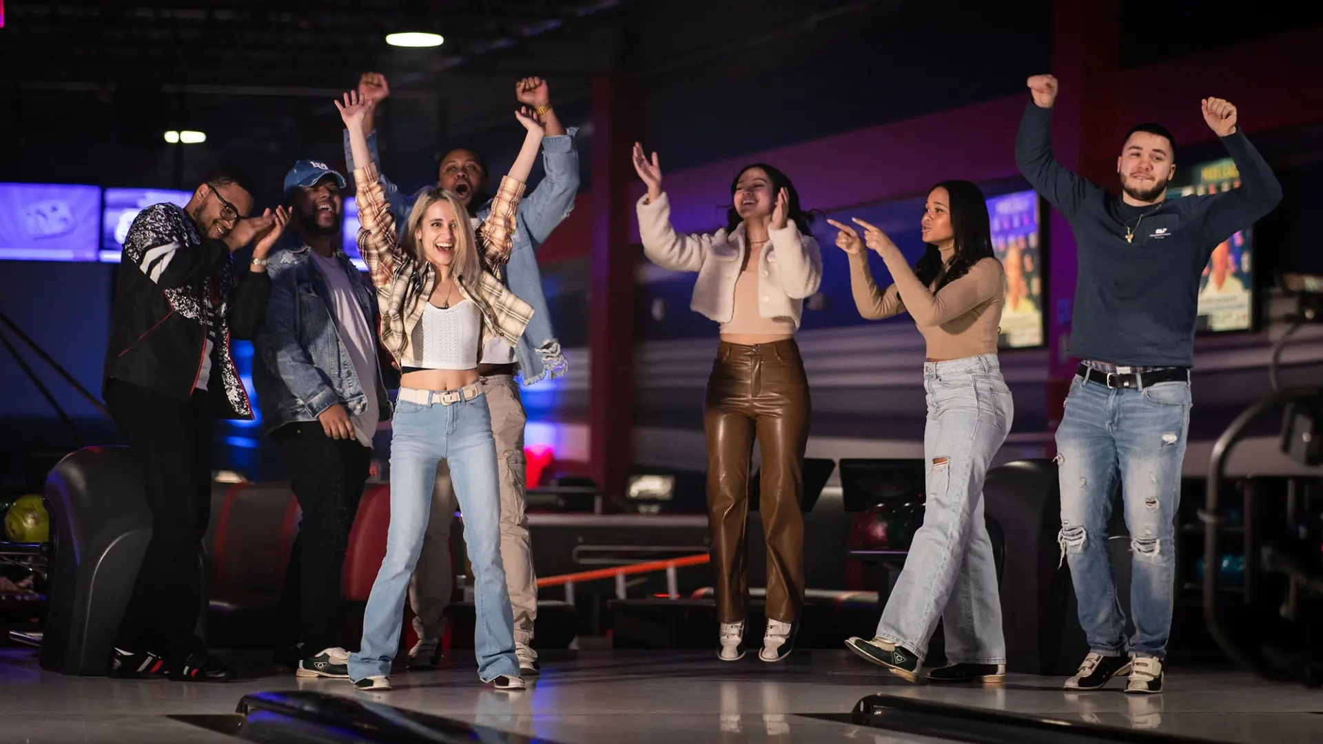 A group of six people is celebrating enthusiastically at a bowling alley called RPM, raising their arms and cheering. They are casually dressed and appear to be having a great time. The background includes bowling lanes and a dimly lit interior with colorful lights.