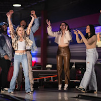 A group of six people is celebrating enthusiastically at a bowling alley called RPM, raising their arms and cheering. They are casually dressed and appear to be having a great time. The background includes bowling lanes and a dimly lit interior with colorful lights.