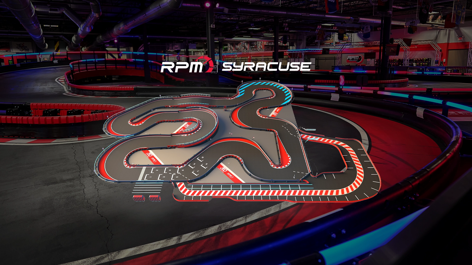 An indoor go-kart track featuring a complex and winding layout with multiple tight turns and straightaways. The track has red, white, and blue markings, with safety barriers along the edges. The surrounding area is dimly lit with a few visible structures.