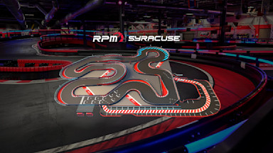 An indoor go-kart track featuring a complex and winding layout with multiple tight turns and straightaways. The track has red, white, and blue markings, with safety barriers along the edges. The surrounding area is dimly lit with a few visible structures.