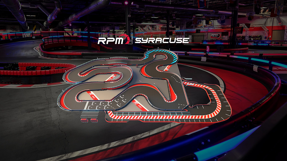 An indoor go-kart track featuring a complex and winding layout with multiple tight turns and straightaways. The track has red, white, and blue markings, with safety barriers along the edges. The surrounding area is dimly lit with a few visible structures.