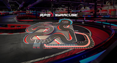 An indoor go-kart track featuring a complex and winding layout with multiple tight turns and straightaways. The track has red, white, and blue markings, with safety barriers along the edges. The surrounding area is dimly lit with a few visible structures.