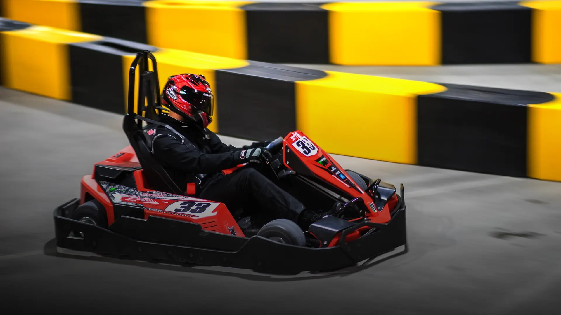 A person wearing a black racing suit and a red helmet is driving a red and black go-kart on an indoor track. The track features a curved section with yellow and black barriers in the background. The driver is focused and navigating the turn.