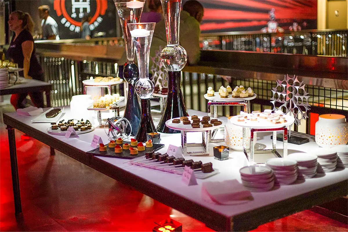 A dessert buffet table is set with an array of pastries, cakes, and sweets on various white and silver stands at the go karting venue. Decorative vases and minimalist sculptures add elegance to the setup. Plates and utensils are neatly arranged, perfect for a post-race celebration, while a person is seen in the background.