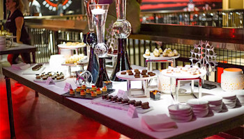 A dessert buffet table is set with an array of pastries, cakes, and sweets on various white and silver stands at the go karting venue. Decorative vases and minimalist sculptures add elegance to the setup. Plates and utensils are neatly arranged, perfect for a post-race celebration, while a person is seen in the background.