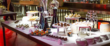 A dessert buffet table is set with an array of pastries, cakes, and sweets on various white and silver stands at the go karting venue. Decorative vases and minimalist sculptures add elegance to the setup. Plates and utensils are neatly arranged, perfect for a post-race celebration, while a person is seen in the background.