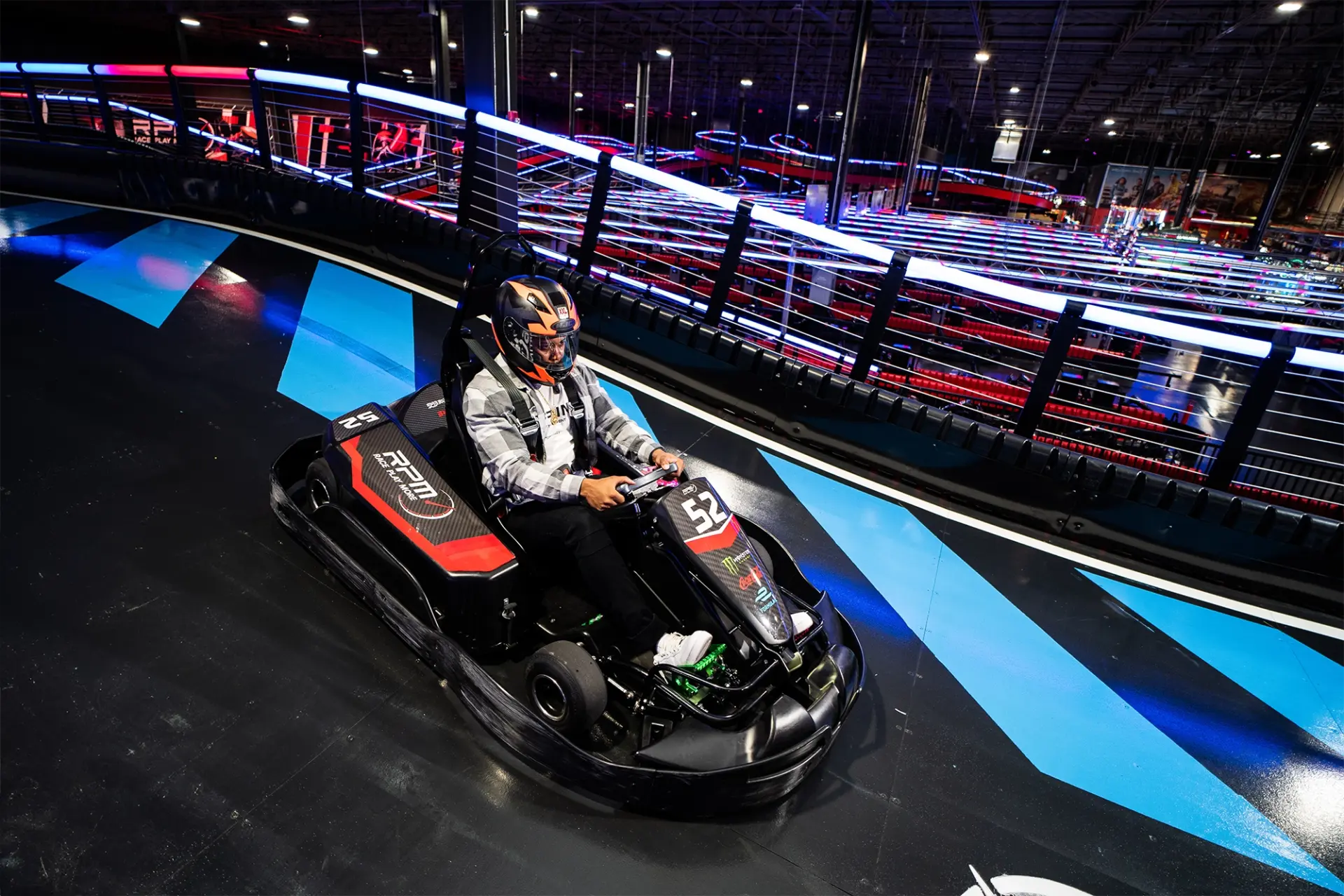 A person wearing a helmet is driving a go-kart on an indoor race track with vibrant blue and white lines. The track features protective barriers and is well-lit with various neon lights in the background.