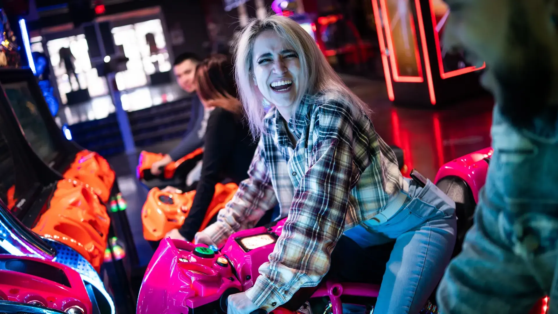 A person with long, light hair smiles broadly while riding an arcade motorcycle game in a neon-lit setting. They are wearing a plaid shirt and jeans. Other individuals are also seen in the background enjoying the games.