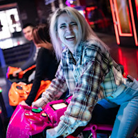 A person with long, light hair smiles broadly while riding an arcade motorcycle game in a neon-lit setting. They are wearing a plaid shirt and jeans. Other individuals are also seen in the background enjoying the games.
