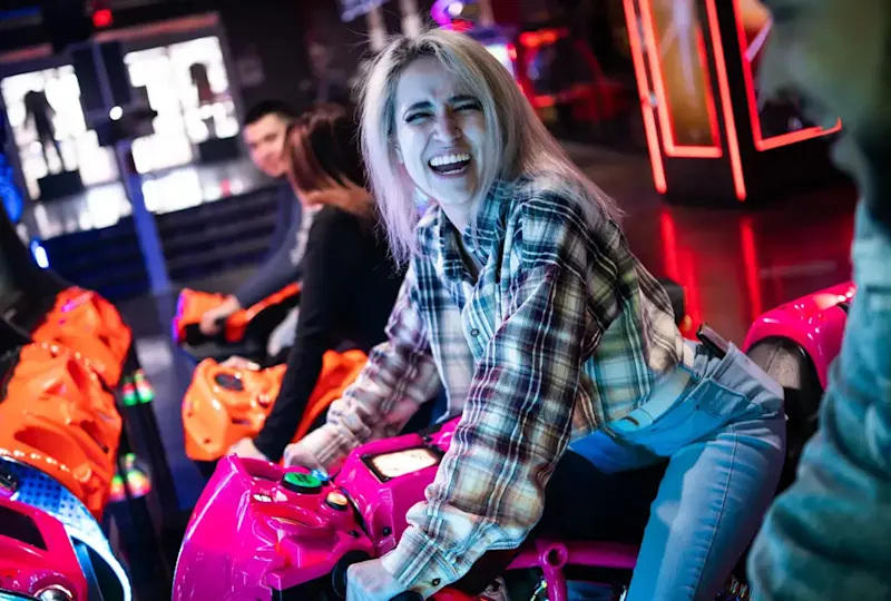 A person with long, light hair smiles broadly while riding an arcade motorcycle game in a neon-lit setting. They are wearing a plaid shirt and jeans. Other individuals are also seen in the background enjoying the games.