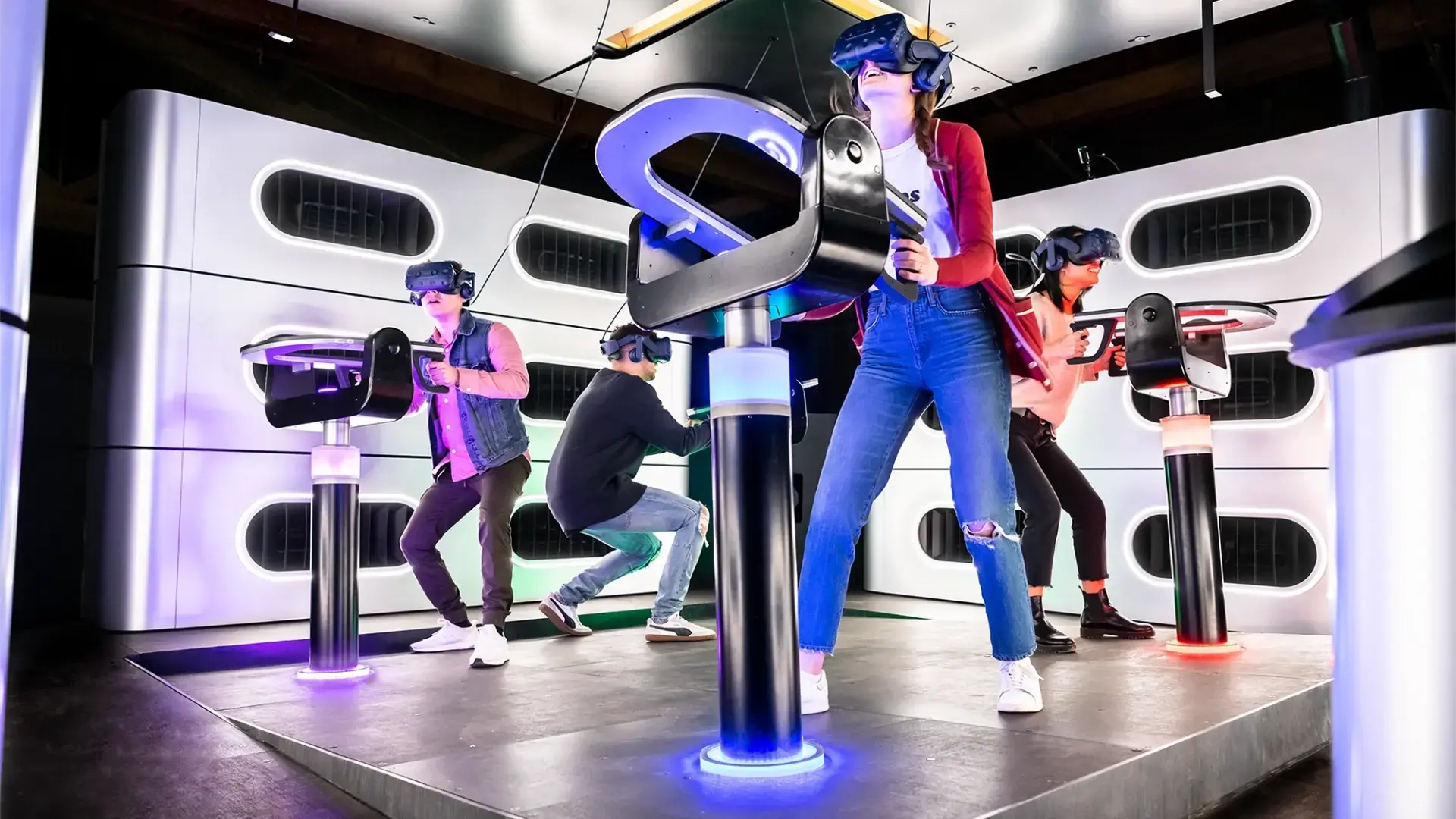 A group of people, wearing VR headsets and holding controllers, are standing on a platform engaging in a virtual reality experience. The room is illuminated with colorful lights and has a futuristic design, including modern black and white decor elements.