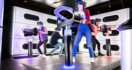 A group of people, wearing VR headsets and holding controllers, are standing on a platform engaging in a virtual reality experience. The room is illuminated with colorful lights and has a futuristic design, including modern black and white decor elements.