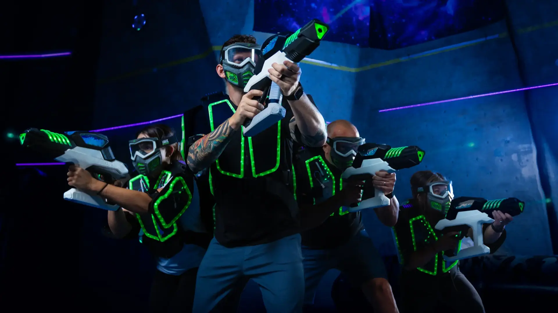 A group of people dressed in futuristic, glowing vests and goggles are engaged in an intense laser tag game, wielding laser guns. They are positioned in an action stance in a dimly lit, neon-lit arena.