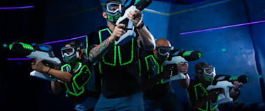 A group of people dressed in futuristic, glowing vests and goggles are engaged in an intense laser tag game, wielding laser guns. They are positioned in an action stance in a dimly lit, neon-lit arena.