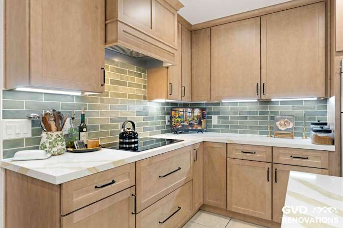 L-shaped maple kitchen with an electric cooktop, quartz counters, sage green subway tile backsplash and under-cabinet lighting
