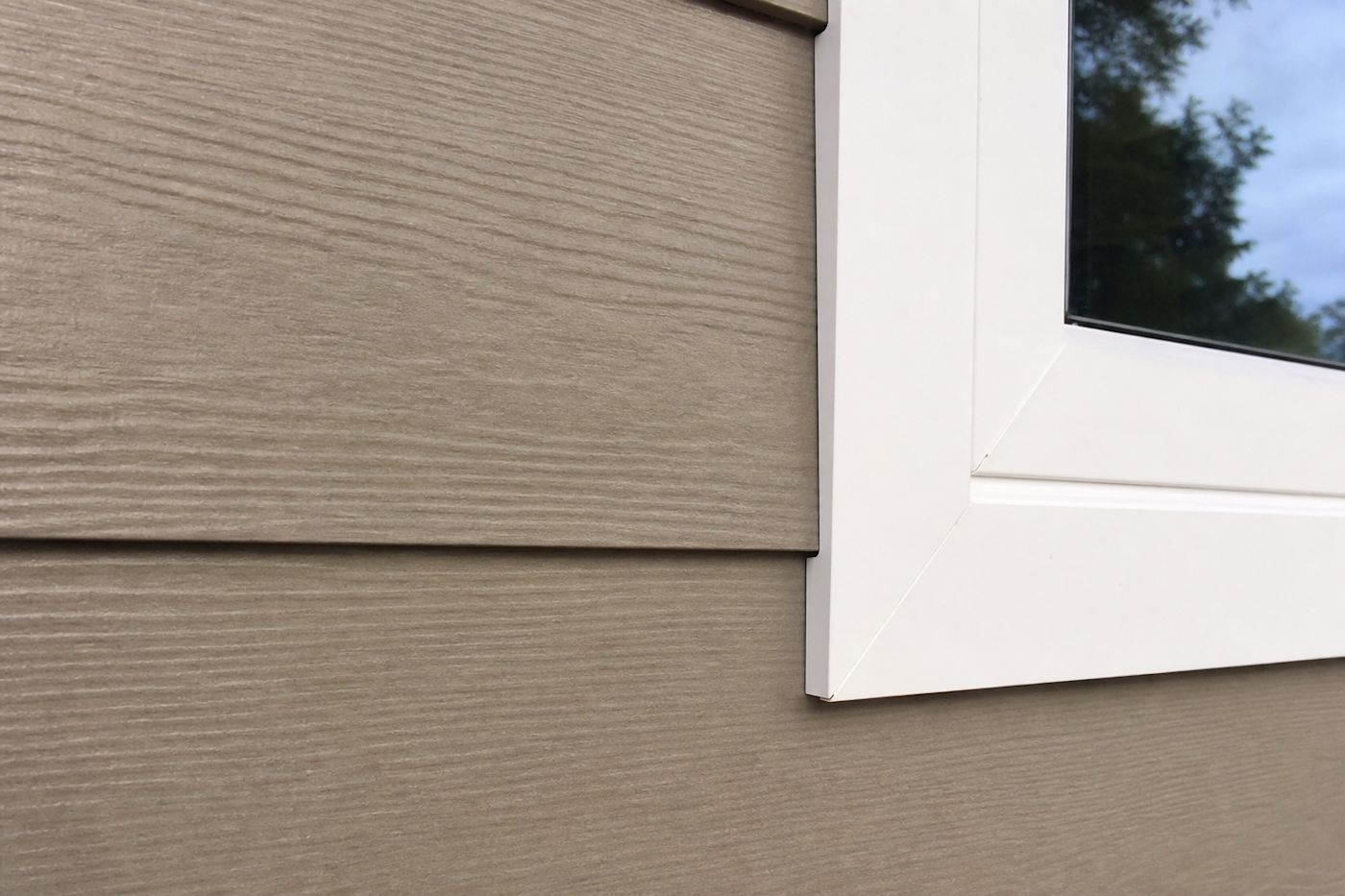 Close-up of warm greige engineered wood lap siding showing woodgrain texture, a clean butt joint, and white window trim