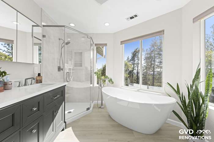 Primary bath with a charcoal double vanity, freestanding tub and a marble-look neo-angle shower by the windows