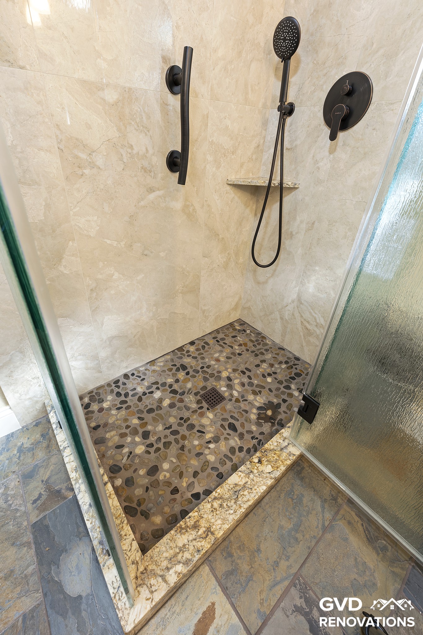 Walk-in shower interior with travertine-look walls, a pebble-tile floor and a grab bar