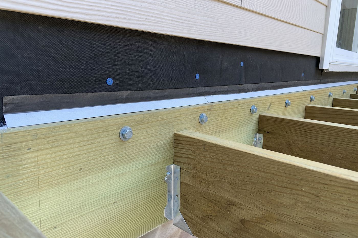 Close-up of a deck ledger board lag-bolted to the house rim joist with metal flashing behind the siding and joist hangers