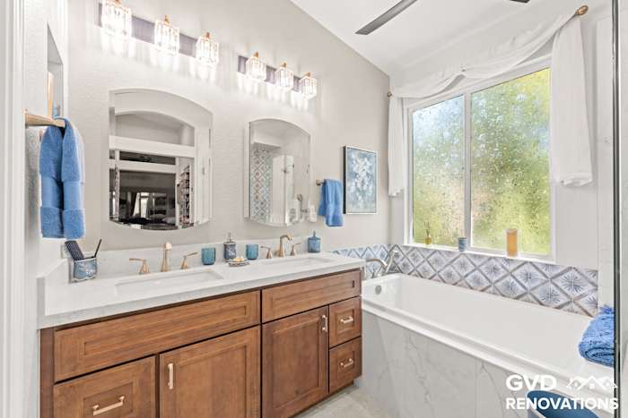 Bathroom vanity and drop-in tub beneath a window with marble-look tile and a blue accent band
