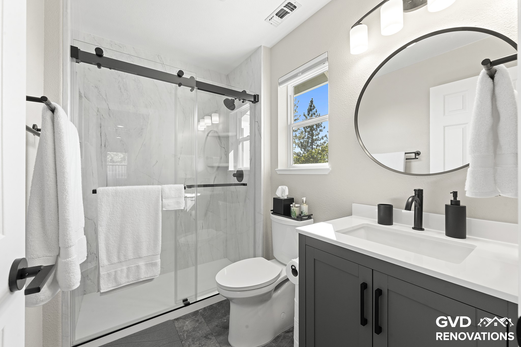 Remodeled hall bathroom in Greenwood with a grey vanity, a round black-framed mirror and a marble-look walk-in shower with a sliding glass door