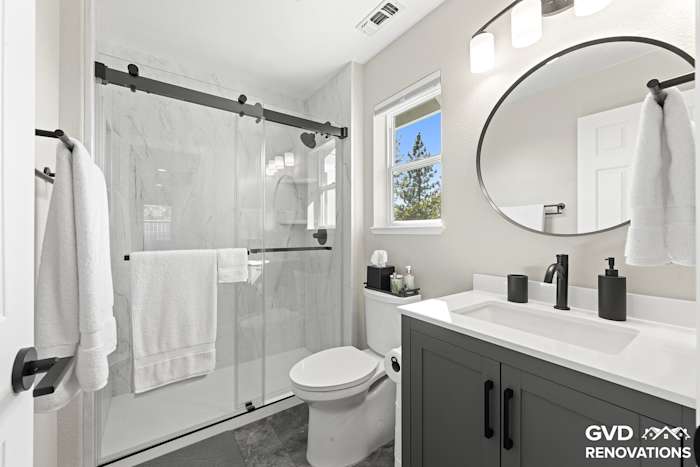 Remodeled hall bathroom in Greenwood with a grey vanity, a round black-framed mirror and a marble-look walk-in shower with a sliding glass door