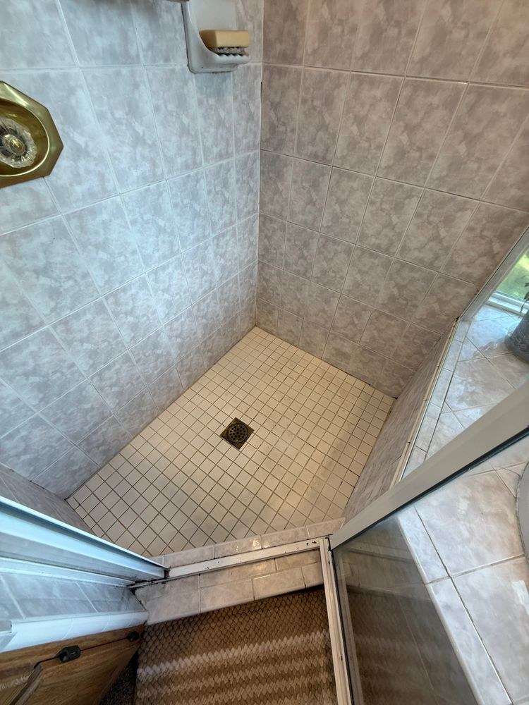 Before: original corner shower stall with grey marble-look tile and a white tile floor